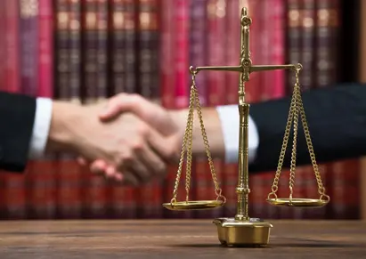 Justice scale on wooden table with judge and client shaking hands in background at courtroom